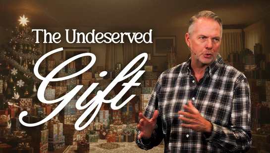A man in a plaid shirt speaks in a room with a Christmas tree and many presents. White text overlay reads, 'The Undeserved Gift'.
